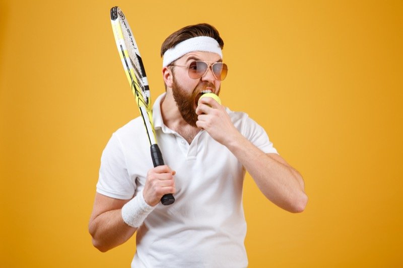 Funny tennis player holding racket and biting ball Man in tennis attire biting a tennis ball against a yellow background