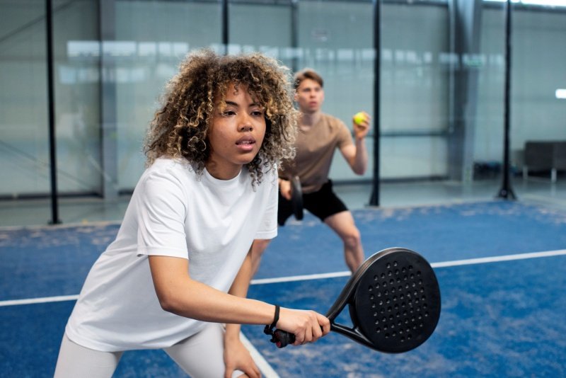 Two people playing padel tennis indoors, one preparing to hit