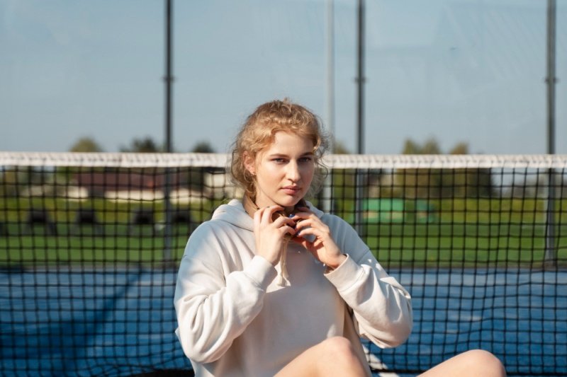 Female padel player in hoodie sitting by the net on court