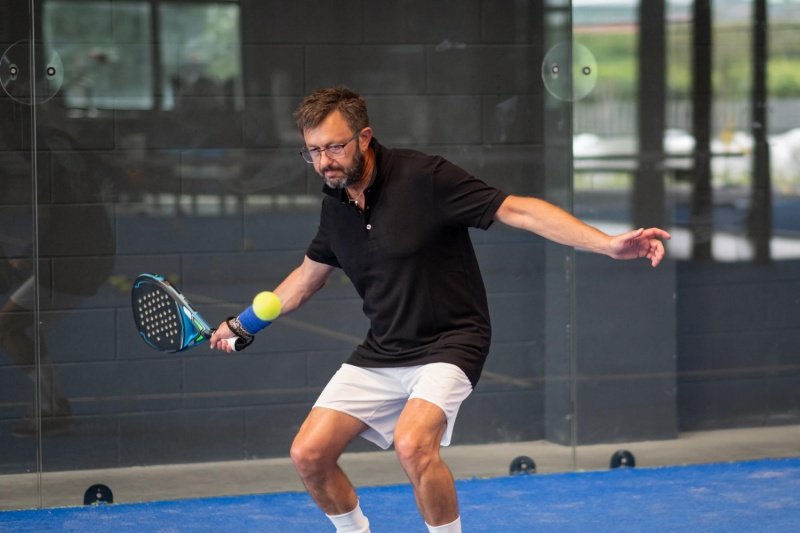 Older male padel player hitting low forehand shot indoors