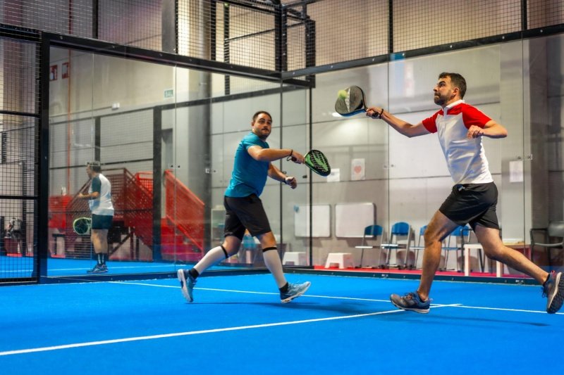Indoor Padel Match two men playing padel indoors on blue court with glass walls